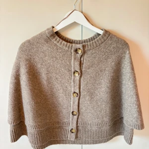 Beige kofta med knappar från Zara - Supermysig beige kofta från Zara med stora knappar framtill. Koftan är stickad med ribbade detaljer vid hals och nederkant, perfekt för lager-på-lager. Avslappnad passform och enkel att styla till olika outfits.