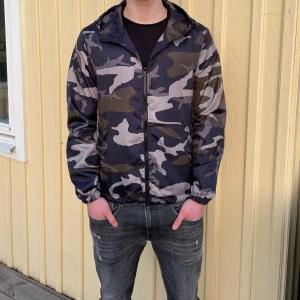 Camouflage vindjacka från Lindfors - Säljer en snygg vindjacka från Lindfors med camouflagemönster i mörkblått, grått och grönt. Jackan har huva, . Perfekt för dig som vill ha en lätt och stilren jacka. Har i alla storlekar och är helt nya och inplastade