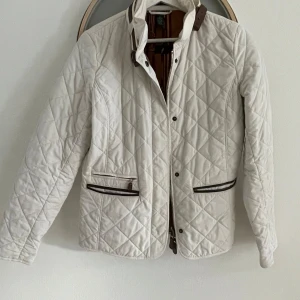 Vit quiltad jacka från Ralph Lauren, storlek S - Snygg vit quiltad jacka från Lauren Ralph Lauren med bruna detaljer vid fickor och krage. Jackan har dragkedja och knappar framtill, samt två fickor med dragkedja. Yttertyg i polyester, fodrad med bomull och polyester. Perfekt för kyliga dagar.