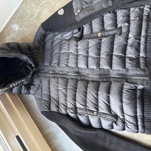 Svart Moncler cardigan - Svart pufferjacka från Moncler med stickade ärmar och rygg, quiltad framsida och huva med dragsko. Jackan har dragkedja framtill, fickor med tryckknappar och en extra bröstficka med dragkedja. Perfekt för dig som vill ha en stilren och varm jacka.