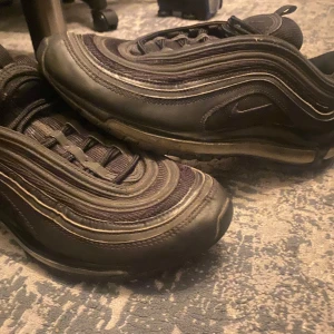 Nike Air Max 97 svarta sneakers - Svarta Nike Air Max 97 sneakers med ikoniska vågformade linjer och synlig Air-sula. Skorna har snörning och är tillverkade i en mix av syntet och textil. Snygg, sportig design med reflekterande detaljer och rund tå. Perfekta för dig som gillar streetwear och klassiska Nike-vibbar. små fel på dom men lite lim kan fixa  Köpt för ett tag sen men nu har dom blivit små   Bra pris vid snabb affär tveka inte fråga frågor