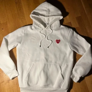 Comme des Garçons Play hoddie - Säljer en vit hoodie från Comme des Garçons Play med det ikoniska röda hjärtat med ögon broderat på bröstet. Tröjan har huva med snörning, magficka och ribbade muddar. Perfekt för dig som gillar stilrena streetwear-plagg. Använd 2 gånger, strl L men sitter som M