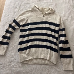 Randig ribbstickad tröja med half zip - Snygg ribbstickad tröja i beige och marinblått med breda horisontella ränder. Tröjan har lång ärm, hög krage och en praktisk half zip-dragkedja framtill. Perfekt för dig som gillar en avslappnad men trendig stil.