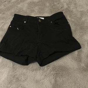 Svarta midwaist shorts i jeans/bomull - Snygga svarta shorts med hög midja och klassisk femficksdesign. De har uppvikta benslut och stängs med knapp och dragkedja. Perfekta för varma dagar och enkla att matcha med allt. Materialet känns mjukt och är troligtvis bomull.