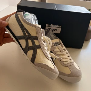 Onitsuka Tiger Mexico 66 sneakers beige - Snygga Onitsuka Tiger Mexico 66 sneakers i beige och grått med klassiska ränder på sidorna. Ovandel i skinn och mocka, vita snören och platt sula med mönstrad undersida. Perfekta för dig som gillar retrostil och vill ha något stilrent till vardags. 