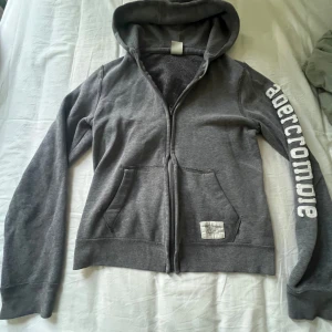 Grå hoodie från Abercrombie - Grå hoodie från Abercrombie med vit logga längs ena ärmen. Tröjan har huva, dragkedja framtill och två fickor. Perfekt för en chill och avslappnad stil.