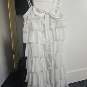 Vit volangklänning med rosett - Superfin vit klänning med tunna axelband, flera lager volanger och en stor rosett framtill. Klänningen har en luftig känsla och är perfekt för sommarens alla tillfällen. Materialet känns lätt och flowy, och volangerna ger en extra söt vibe.