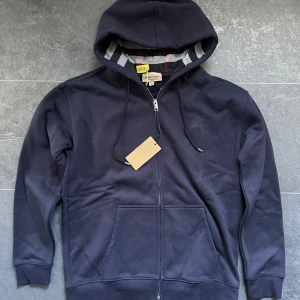 Mörkblå hoodie från Burberry M - Säljer en mörkblå hoodie från Burberry med dragkedja, huva med snörning och klassiskt rutigt foder. Tröjan har broderad logga på bröstet och stora fickor framtill. Perfekt för dig som gillar stilrena och exklusiva plagg. I ny sick och är en perfekt tröja för kallare dagar då den är väldigt tjock. 
