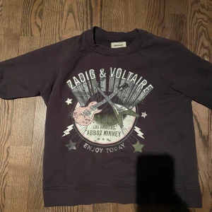 Svart sweatshirt Zadig & Voltaire - Cool svart sweatshirt från Zadig & Voltaire med stort tryck framtill med gitarrer, stjärnor och texten 'Los Angeles Abbot Kinney'. Tröjan har rund hals och långa ärmar. Perfekt för dig som gillar rockig stil.