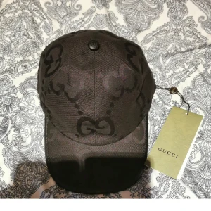 Gucci Keps - Snygg svart keps från Gucci med det klassiska GG-logomönstret i mörk ton. Kepsen har böjd skärm och justerbar rem baktill. Tillverkad i Italien och kommer med originaltaggar. Perfekt för dig som vill ha en lyxig och trendig accessoar.