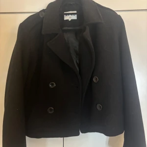 Svart skepparkavaj från NA-KD, stl 34 - Stilren svart skepparkavaj från NA-KD i klassisk modell med dubbelknäppning och axelklaffar. Jackan är tillverkad i ullblandning och har en rak, något croppad passform. Perfekt för dig som vill ha en tidlös och snygg look.