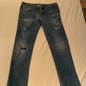 Blå slitna jeans från dondup, stl 31 - Säljer ett par blå jeans från dondup George i storlek 31. Jeansen har flera slitningar och hål framtill för en riktigt trendig look. Klassisk femficksmodell med knappgylf och smal passform. Perfekta för dig som gillar en avslappnad och edgy stil.