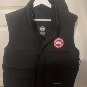 Svart dunväst från Canada Goose - Svart dunväst från Canada Goose med klassisk logga på bröstet och praktiska fickor framtill. Västen har hög krage och stängs med både dragkedja och knappar. Perfekt för lager-på-lager och riktigt snygg till streetwear. Använd max 5 gånger