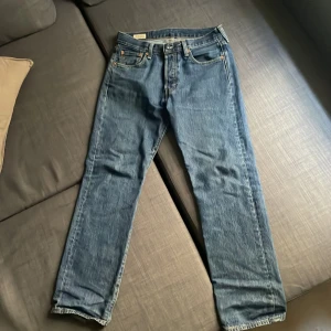 Levi's 501 blå jeans W30 L30 - Klassiska blå Levi's 501 jeans med raka ben och femficksmodell. Jeansen har en snygg mellanblå tvätt, knäppning med knappar och detaljer i kopparfärgad metall. Tillverkade i slitstarkt bomullsjeans. Perfekta för en avslappnad och tidlös look.