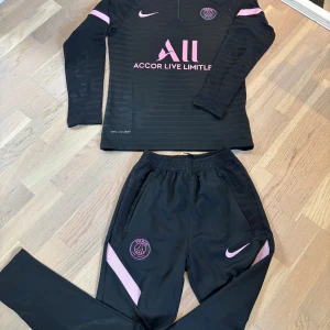 Svart PSG fotbollströja från Nike - Säljer en svart långärmad PSG fotbollströja från Nike med rosa detaljer och klubbmärke på bröstet. Tröjan har halv dragkedja och är tillverkad i Dri-FIT material som andas. Snyggt tryck med Accor Live Limitless på framsidan och diskreta ränder i tyget.