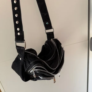 Svart manchesterväska med dragkedjor - Cool svart axelväska i manchester med bred justerbar rem och flera silvriga dragkedjor. Väskan har flera fack och detaljer i metall, perfekt för dig som gillar en edgy och unik stil. Rymmer allt du behöver till vardagen.