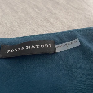 Blå oneshoulder jumpsuit Josie Natori - Snygg blå jumpsuit  från Josie Natori i oneshoulder-modell med lång ärm och coola volangdetaljer vid midjan. Materialet är mjukt och har ett elegant fall, perfekt för dig som vill sticka ut med en unik siluett och asymmetrisk design.