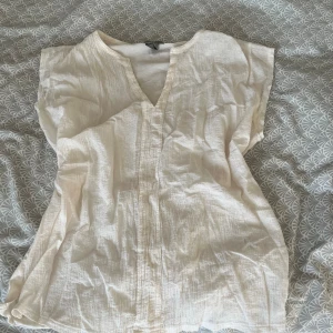 Vit blus med v-ringning från Lindex - Luftig och lätt vit blus från Lindex i storlek S. Blusen har v-ringning, korta ärmar och är tillverkad i ett krinklat bomullstyg som ger en avslappnad känsla. Perfekt för varma dagar och enkel att styla till olika looks. Hör av dig vid frågor💗