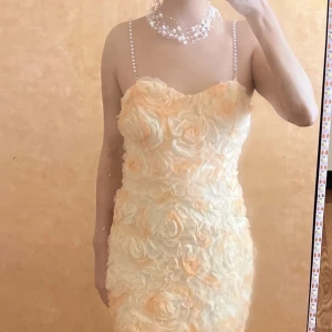 Champagnefärgad blommig kortklänning och 2 present  - klänning i champagne och vitt med 3D-blommor över hela tyget. Klänningen har pärlband som axelband och en söt, figurnära form. Materialet är lätt och har en satinliknande insida. Dragkedja bak för enkel påklädning.  Present (1 halsband i bild och 1 mask)