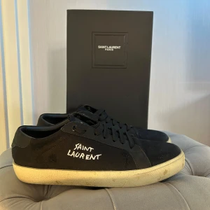Saint Laurent skor - Svarta sneakers från Saint Laurent med vit broderad logga på sidan. Skorna har snörning, platt sula i vitt och rund tå. Medföljer originalkartong och dustbag samt allt annat og. Strl 42. Säljes då jag inte använder dom längre. Bra skick, endast lite smutsiga! Mvh