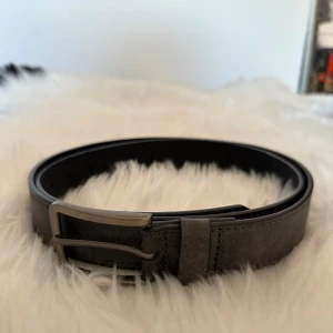 Mörkgrå mockabälte - Snyggt mörkgrått bälte i mocka med stilrent metallspänne. Bältet har en klassisk, rak form och är perfekt för att ge din outfit en clean och modern touch. Passar till jeans eller kostymbyxor och har diskreta sömmar längs kanterna.