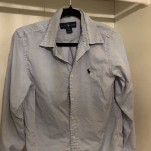 Ljusblå Ralph Lauren skjorta - Klassisk ljusblå skjorta från Ralph Lauren med broderad logga på bröstet. Skjortan har lång ärm, knappar framtill och en snygg krage. Perfekt för dig som gillar stilrena och tidlösa plagg. Storleken är 14 år vilket ungefär motsvarar XS.