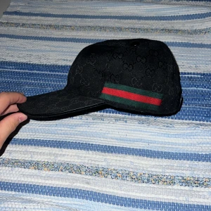 Svart Gucci keps med GG-mönster - Svart keps från Gucci med diskret GG-monogram över hela ytan och klassiska gröna och röda ränder på sidan. Kepsen har böjd skärm och justerbar baksida. Tillverkad i bomull och polyester, perfekt för dig som vill ha en stilren och lyxig accessoar.