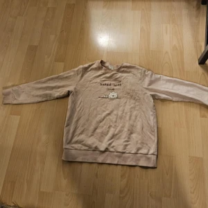 Beige sweatshirt med söt björn från GIADAO - Beige sweatshirt med gulligt broderi av en björn och texten 'baked with love' på bröstet. Tröjan har rund hals, långa ärmar och ribbade muddar. Perfekt för dig som gillar söta detaljer och en avslappnad stil.