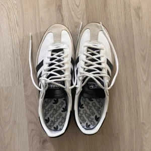 Adidas Handball Spezial vita sneakers - Säljer ett par Adidas Handball Spezial sneakers i vitt med svarta detaljer och klassiska tre ränder på sidorna. I storlek 43 1/3. I nytt skick, utan prislapp. Nytt pris är runt 1000kr. Ovandelen är i skinn och mocka, med vit snörning och gummisula. Snygg retrostil med svart häl och tunga, samt guldfärgad 'Spezial'-text på sidan. Kontakta mig vid frågor eller för fler bilder. 
