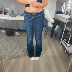 Mörkblå bootcut jeans med kontrastsömmar - Säljer ett par mörkblå jeans med bootcut passform och tydliga vita kontrastsömmar. Jeansen har fickor med lock och knapp bak samt klassisk femficksdesign. Materialet är jeans och modellen har låg midja. Perfekta för dig som gillar en retroinspirerad look.