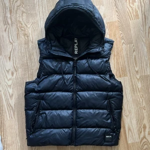 Svart dunväst med huva från Replay - Svart dunväst från Replay med huva och dragkedja framtill. Västen har två fickor med dragkedja och en quiltad design som ger en cool och sportig look. Perfekt för lager-på-lager och funkar till många outfits.