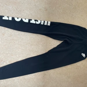 Svarta Nike mjukisbyxor med tryck - Svarta mjukisbyxor från Nike med stort vitt 'JUST DO IT.'-tryck längs ena benet och Nike-logga vid höften. Byxorna har elastisk midja och är tillverkade i mjukt bomullsmaterial. Perfekta för chill eller träning. S men sitter väldigt tight, passar även XS