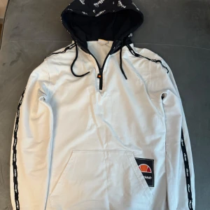 Vit hoodie från Ellesse med svart huva - Vit hoodie från Ellesse med svart huva och logga, samt svarta detaljer längs ärmarna. Halv dragkedja framtill, känguruficka och snörning i huvan. Perfekt för en chill och sportig look.