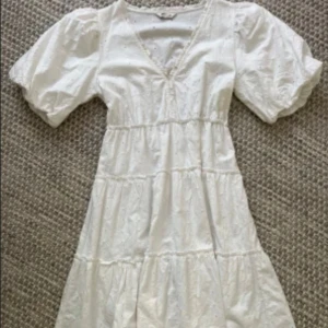 Vit klänning med puffärm - Säljer en vit klänning med v-ringning och omlottdetalj. Klänningen har puffiga korta ärmar och volanger längs kjolen. Tillverkad i bomull med broderade detaljer som ger en romantisk känsla. Perfekt för sommarens alla tillfällen!