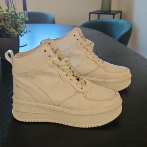 Vita höga sneakers med platåsula - Säljer ett par vita höga sneakers med rejäl platåsula och snörning. Skorna har rund tå, perforerade detaljer på tån och är tillverkade i syntetmaterial. Perfekta för dig som vill ha en clean och modern look med extra höjd.