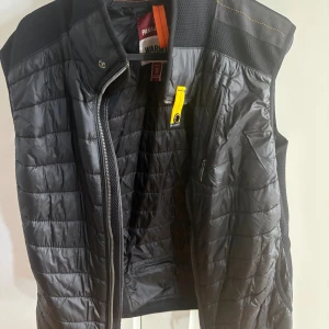 Svart dunväst från Parajumpers - Snygg svart dunväst från Parajumpers med quiltad design och gul detalj vid dragkedjan. Västen har hög krage, dragkedja framtill och en bröstficka med dragkedja. Perfekt för lager-på-lager och streetstyle.