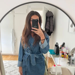 Denim jacka med knytband (slutsåld i butik) - Säljer en snygg  oanvänd denim jacka som går över rumpan i blå denim med knytband i midjan och stora fickor framtill. Jackan har långa ärmar med uppvikta muddar och omlottstängning. Perfekt för dig som gillar en avslappnad men trendig och elegant look. (Jackan är helt slutsåld i butik)