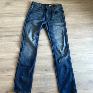 Blå jeans från Wrangler med raka ben - Snygga blå jeans från Wrangler med klassisk femficksdesign och raka ben. Jeansen har tydliga kontrastsömmar, läderpatch med logga på bakfickan och slitningar framtill för en cool look. Perfekta för dig som gillar en tidlös och avslappnad stil.