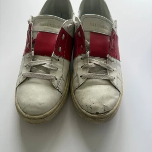 Valentino Garavani Open sneakers vit/röd - Snygga Valentino Garavani Open sneakers i vitt skinn med bred röd läderdetalj över snörningen. Klassisk rund tå, platt sula och ikoniska nitar baktill. Perfekta för dig som vill ha en statement-sneaker med lyxig känsla och cool design.