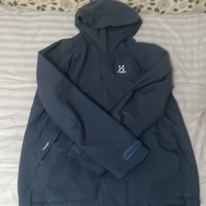 Mörkblå vindjacka från Haglöfs - Säljer en mörkblå vindjacka från Haglöfs med huva och halv dragkedja. Jackan har minimalistisk design, meshfoder på insidan och diskret logga på bröstet. Perfekt för blåsiga dagar och outdoor-äventyr.
