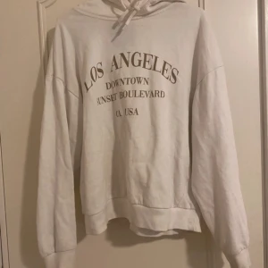 Vit hoodie med Los Angeles-tryck - Vit hoodie med tryck i brunt framtill. Tröjan har huva med snörning och långa ärmar. Perfekt för en chill och avslappnad stil.