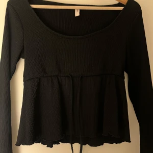 Svart långärmad blus från Nelly - Säljer en svart långärmad blus från Nelly med rundad halsringning och smockad struktur. Toppen har en volangkant nertill och knytband i midjan för en snygg passform. Perfekt att styla med jeans eller kjol.