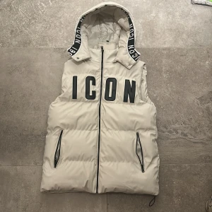 Beige dunväst med huva och tryck - Stilren beige dunväst med stor svart 'ICON'-text framtill och huva med svartvit textdetalj. Västen har dragkedja framtill, två fickor med dragkedja och justerbar dragsko nedtill. Perfekt för lager-på-lager och streetstyle.