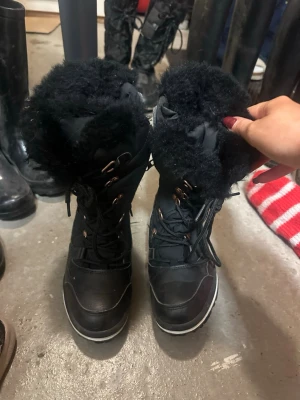 Svarta vinterboots med fuskpäls - Säljer ett par svarta vinterboots med snörning och fluffig fuskpäls runt skaftet. Skorna har grov sula för bra grepp och detaljer i metall. Perfekta för kalla dagar och riktigt snygga till vinterlooken.
