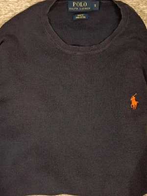 Mörkblå tröja från Polo Ralph Lauren - Snygg mörkblå tröja från Polo Ralph Lauren i slim fit-modell, tillverkad av mjuk pima bomull. Klassisk rund hals och orange broderad logga på bröstet. Perfekt för dig som gillar stilrena och tidlösa plagg.