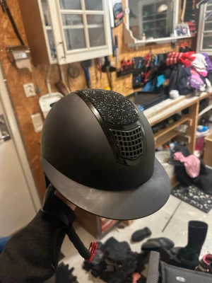 Svart CRW ridhjälm med glitterdetalj - Säljer en svart ridhjälm från CRW i storlek M. Hjälmen har en snygg glitterdetalj på ovansidan och ett ventilerande galler framtill. Justerbart hakband och mjuk, vadderad insida för extra komfort.