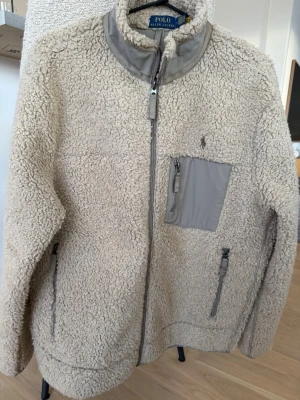 Beige teddyjacka från Polo Ralph Lauren - Säljer en beige teddyjacka från Polo Ralph Lauren med hög krage och dragkedja framtill. Jackan har en grå bröstficka med dragkedja, två sidofickor med dragkedja och broderad logga på bröstet. Perfekt för kalla dagar och riktigt mysig att bära.
