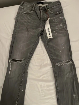 Grå ripped jeans från Purple Brand - Säljer ett par grå jeans från Purple Brand med slitna detaljer och hål vid knäna. Jeansen har en skinny passform och coola färgstänk för en edgy look. Klassisk femficksmodell och soprena sömmar. 