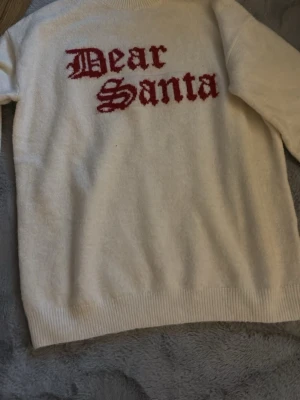 Vit jultröja Dear Santa H&M - Mysig vit stickad tröja från H&M med röd text 'Dear Santa' på bröstet. Perfekt för julen och har en klassisk rund halsringning och långa ärmar. Materialet är mjukt och skönt, passar dig som vill ha en chill julstil.