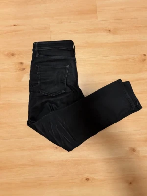 Svarta tvättade jeans från Jack & Jones - Säljer ett par svarta tvättade jeans från Jack & Jones med klassisk femficksdesign och bekväm passform. Jeansen har raka ben och subtila slitningar framtill för en snygg look. Materialet är mjukt och stretchigt för extra komfort.